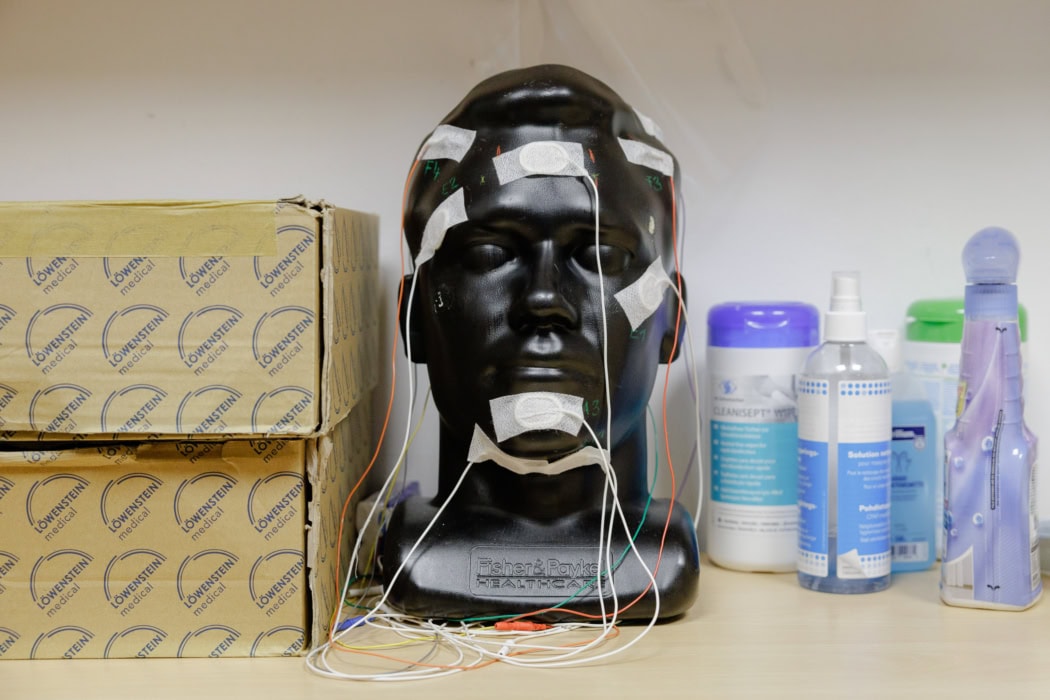 Schwarzer Modellkopf mit Elektroden und Kabeln für medizinische Messungen des EEG (Hirnstrombild), flankiert von Kartons mit „Löwenstein medical“-Aufschrift und diversen Reinigungs- und Desinfektionsmitteln im Hintergrund. Vorbereitung für Tests der Schlafdiagnostik.
