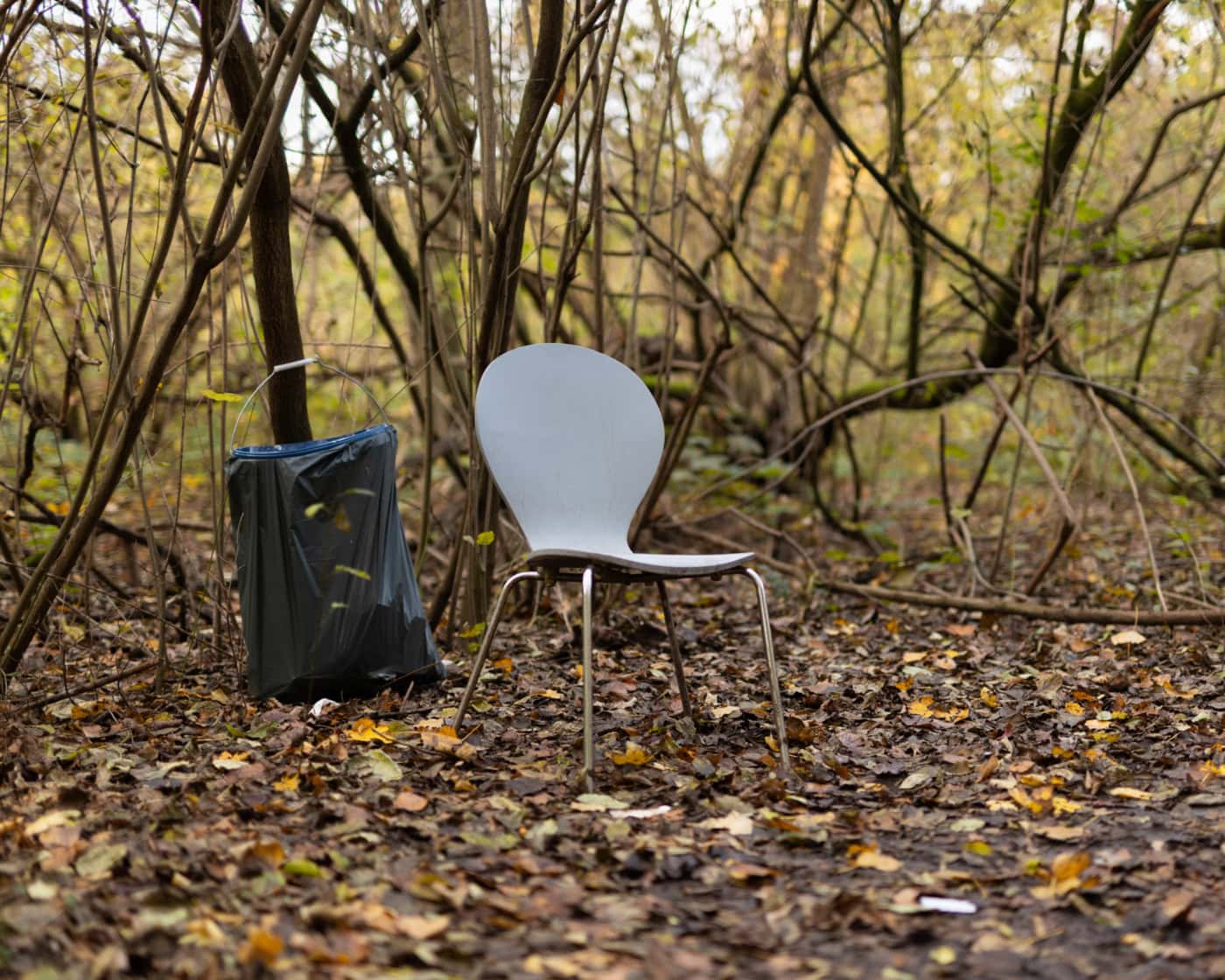 Das Foto ist in einem Waldstück aufgenommen worden. Der Boden ist mit Laub bedeckt und es sind Büsche und kleinere Bäume im Hintergrund erkennbar. Im Zentrum steht ein grauer Stuhl und ein Mülleimer.