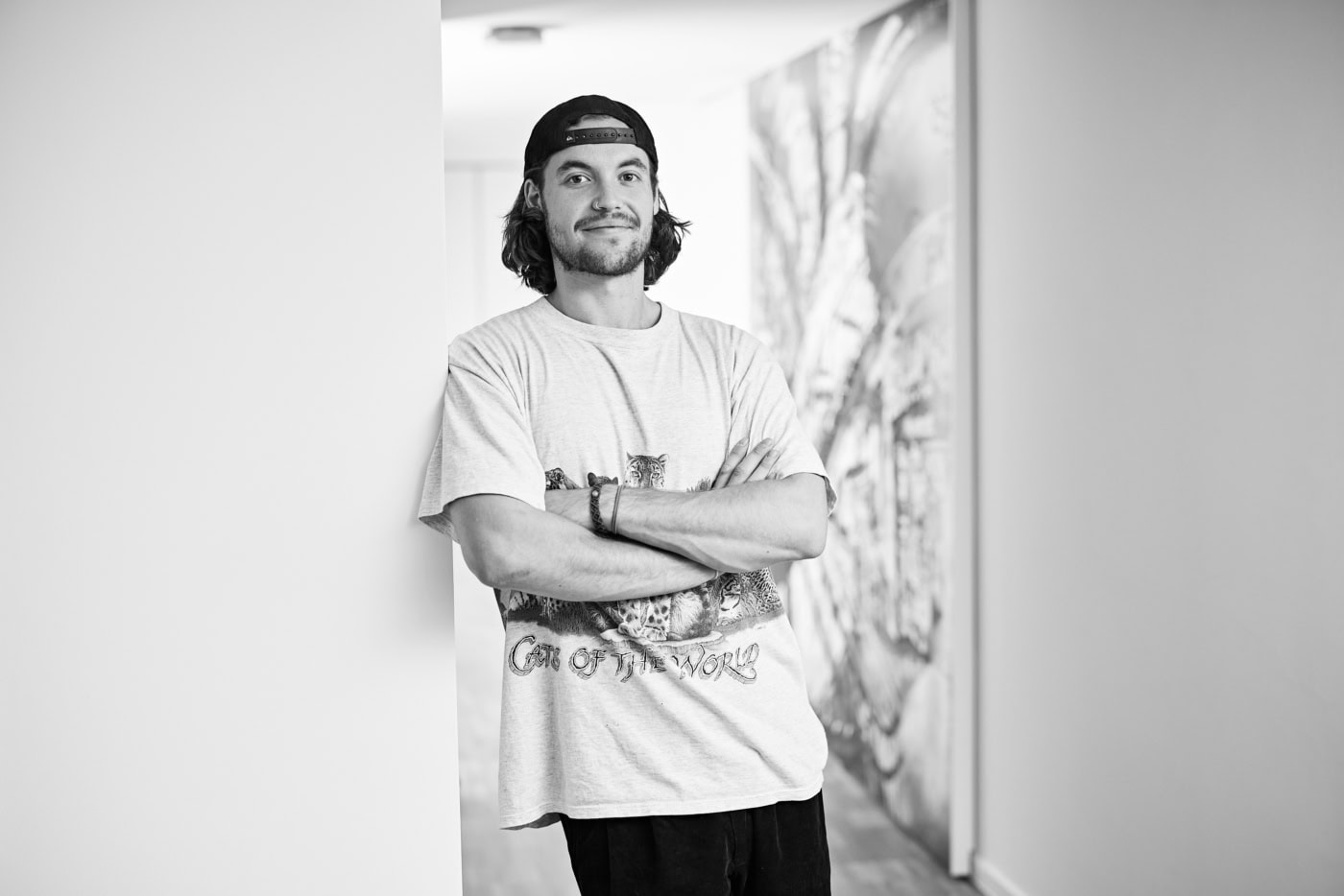 Kilian steht mit an einer Wand und lächelt in die Kamera. Seine Arme schränkt er vor sich ein. Er trägt ein weißes Tshirt und eine Cap. Er hat kinnlange Haare und ein Nasenpiercing.