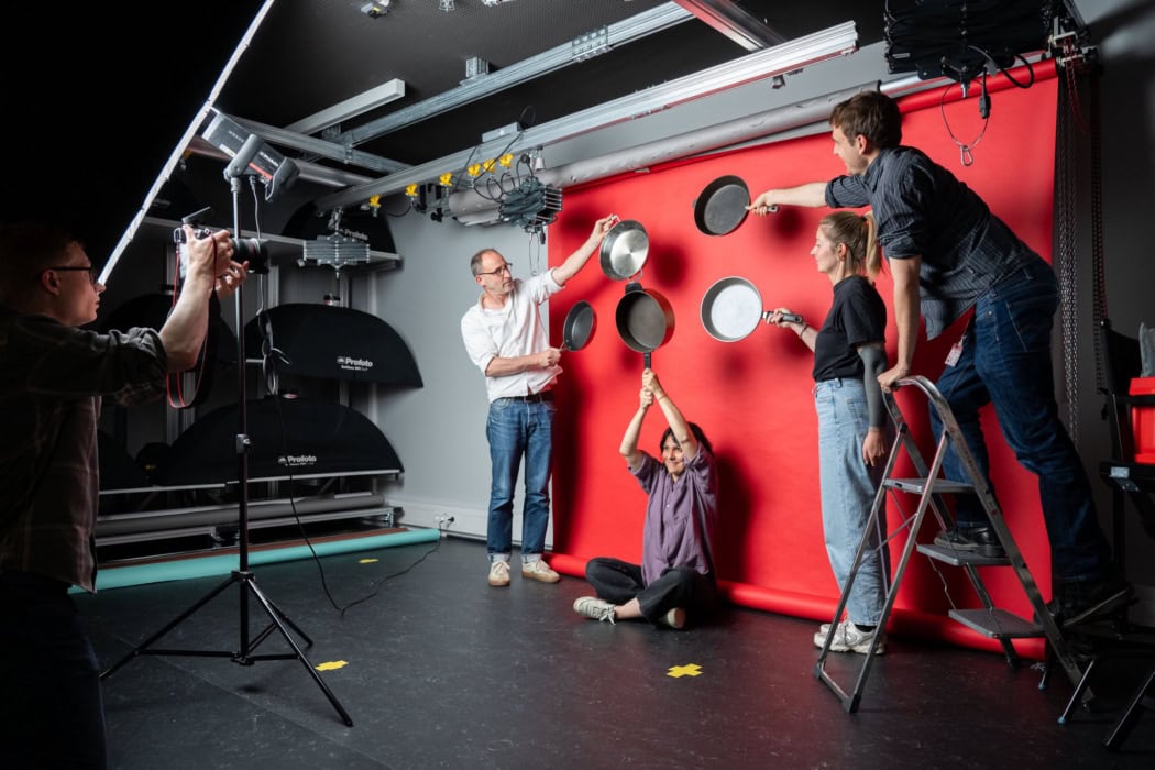 Fünf Personen im Fotostudio der F.A.Z. inszenieren mit Bratpfannen eine kreative Szene. Im Mittelpunkt: Sophie Boyer und Stefan Nieland, beide im journalistischen Praktikum aus dem Studiengang Visual Journalism and Documentary Photography in Hannover. Die Szene zeigt Zusammenarbeit im Redaktionspraktikum und gestalterische Praxis im Journalismus.