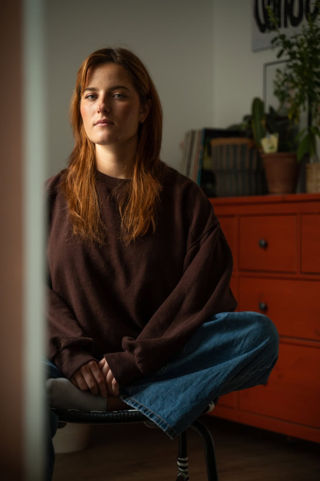 Portrait von Luisa Hannke. Sie sitzt im Schneidersitz auf einem Stuhl in einem Zimmer. Hinter ihr ist eine Kommode zu sehen mit einer Pflanze darauf. Licht durch ein Fenster trifft ihr Gesicht von rechts. Sie blickt in die Kamera. Ihre langen, roten Haare liegen offen über beiden Schultern.