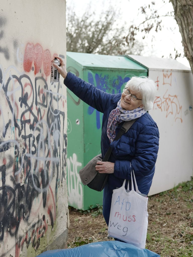 Irmela Mensah-Schramm in blauer Jacke und buntem Schal übersprüht Graffiti an einer Wand und lächelt. Sie trägt eine Tasche mit der handgeschriebenen Botschaft "AfD muss weg".