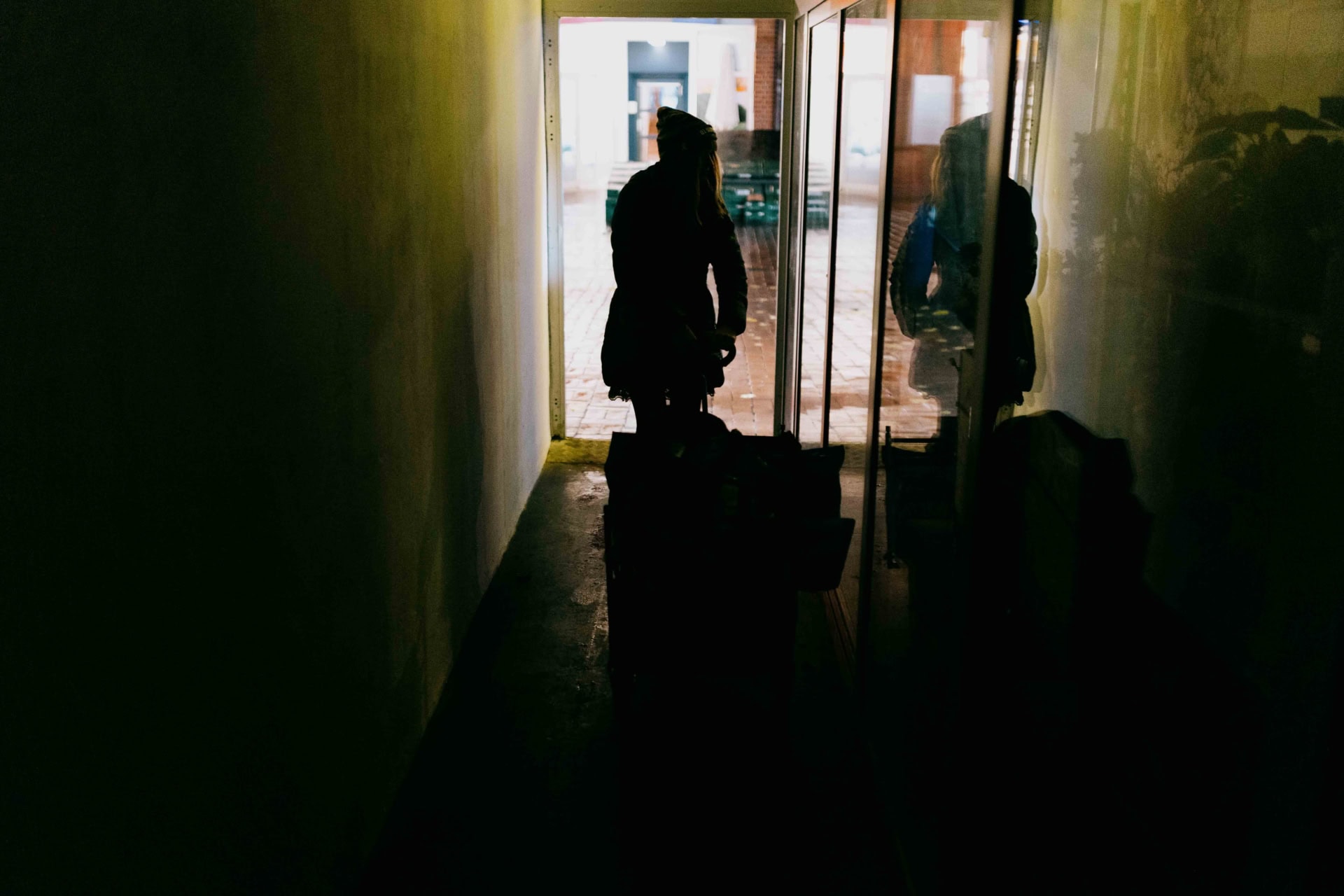 Eine Silhouette einer Person mit Mütze, die einen Wagen durch einen dunklen Gang schiebt. Im Hintergrund ist ein gepflasterter Hof zu sehen, beleuchtet durch Tageslicht, das durch eine Türöffnung einfällt. Die Spiegelung der Person ist auf der Glaswand rechts im Bild sichtbar.