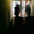 Eine Silhouette einer Person mit Mütze, die einen Wagen durch einen dunklen Gang schiebt. Im Hintergrund ist ein gepflasterter Hof zu sehen, beleuchtet durch Tageslicht, das durch eine Türöffnung einfällt. Die Spiegelung der Person ist auf der Glaswand rechts im Bild sichtbar.
