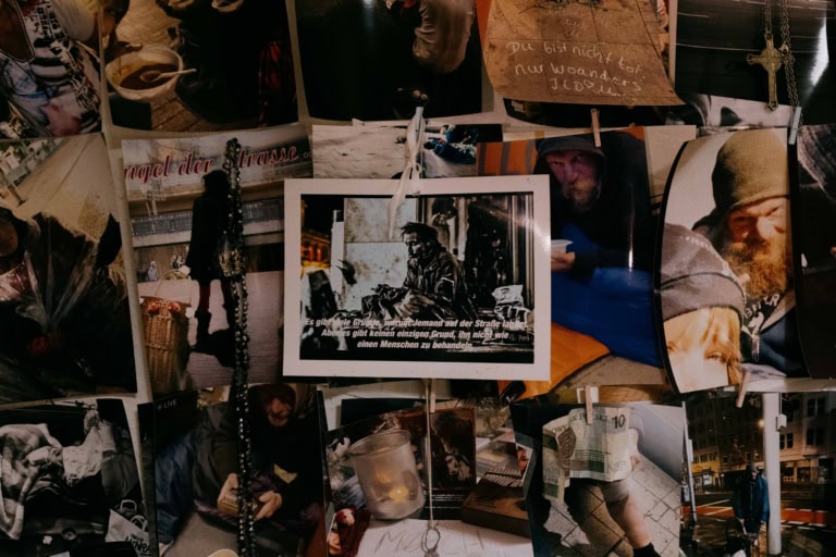 Dieses Bild zeigt eine Collage aus verschiedenen Fotografien und Erinnerungsstücken, die Personen darstellen, die Obdachlosigkeit erleben, handgeschriebene Notizen und persönliche Bilder. Ein zentrales Bild enthält den deutschen Text: „Es gibt viele Gründe, warum jemand auf der Straße landet. Aber es gibt keinen einzigen Grund, ihn nicht wie einen Menschen zu behandeln.“ Die umliegenden Fotos und Objekte spiegeln das Thema der Widerstandskraft und Menschlichkeit in schwierigen Lebenssituationen wider.