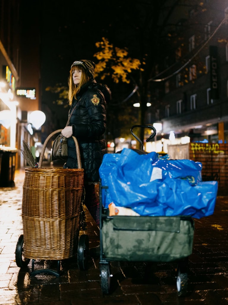 Eine Frau in einer schwarzen Jacke mit Mütze steht auf einer nassen, beleuchteten Straße bei Nacht. Sie hält einen Korbwarenwagen mit Griffen und steht neben einem grünen Bollerwagen, der mit blauen Plastiktüten beladen ist. Im Hintergrund sind Geschäfte, Lichter und herbstliche Bäume zu erkennen.
