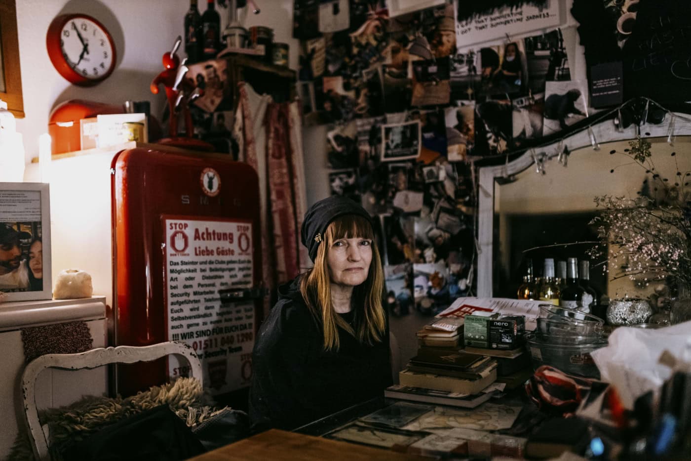 Porträt von Martina "Nachtengel" an einem Tisch in ihrer privaten Wohnung. Im Hintergrund ist ein großer roter Schrank zu sehen und einer Pinnwand mit ihren privaten Fotos.