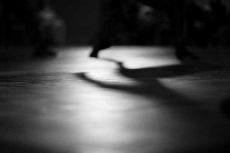 Abstract black-and-white photograph focusing on a shadow cast across the floor, likely from a moving figure. The lighting highlights the blurred edges of the shadow, creating a sense of motion and mystery.