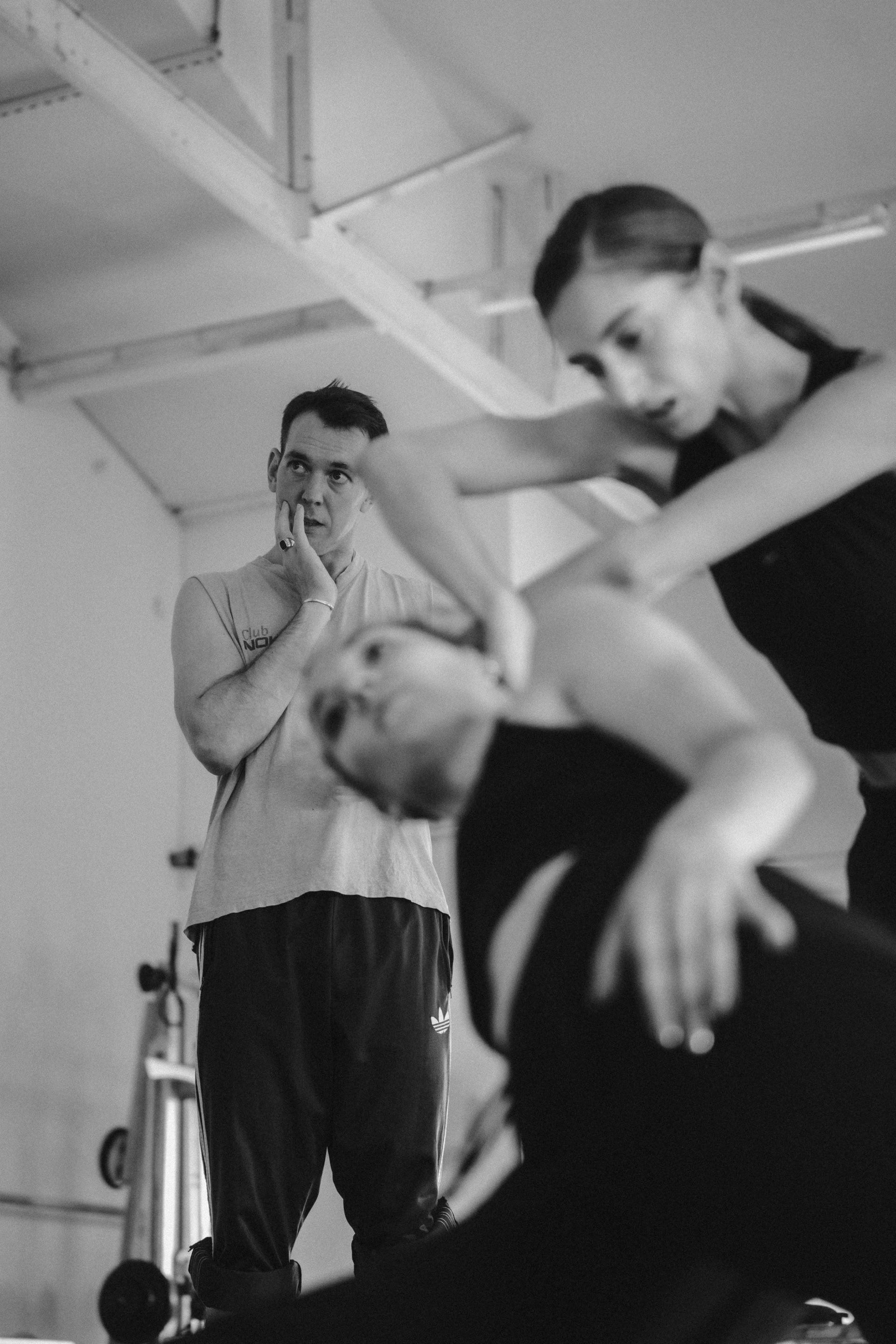 Schwarz-Weiß-Foto eines Mannes, der nachdenklich in einem Tanzstudio steht und zwei Tänzerinnen in Bewegung beobachtet. Eine Tänzerin ist in der Mitte des Bildes scharf abgebildet, während die zweite Tänzerin im Vordergrund unscharf erscheint, was den Eindruck der Bewegung verstärkt. Die Decke im Industriestil und die weiche Beleuchtung verleihen der Szene Tiefe.