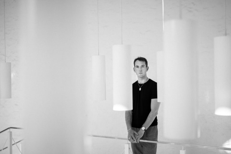 Schwarz-Weiß-Porträt eines Mannes, der in einem minimalistischen, gut beleuchteten Innenraum steht, in dem zylindrische Pendelleuchten um ihn herum hängen. Er trägt ein schwarzes T-Shirt, eine Halskette und eine Uhr und blickt mit ruhigem Blick direkt in die Kamera. Die Komposition betont Licht, Schatten und Symmetrie.