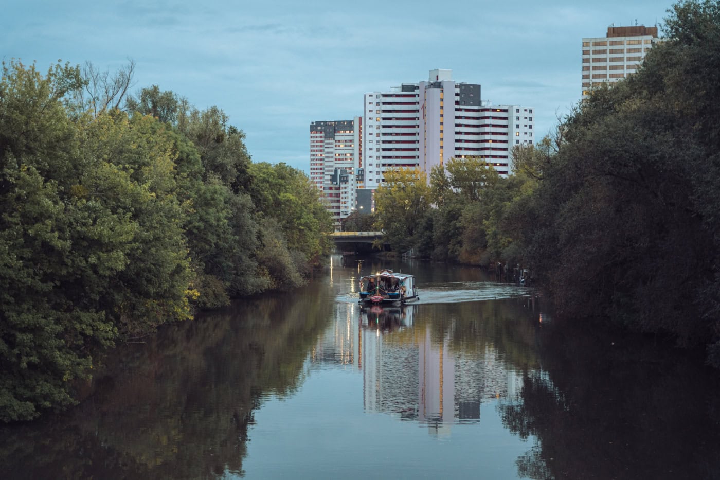 Auf einem ruhigen Fluss, umsäumt von grünen Bäumen ist ein Hausboot zu sehen, das in Richtung des Betrachtenden schwimmt. Im Hintergrund ragt das hannoveraner Hochhaus Ihmezentrum empor.