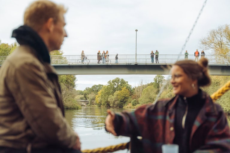 Zwei Personen, die unscharf im Vordergrund zu sehen sind, unterhalten sich. Im Hintergrund scharf zu erkennen: Eine Brücke über dem Fluss, auf dem Personen stehen und in Richtung der Kamera blicken.