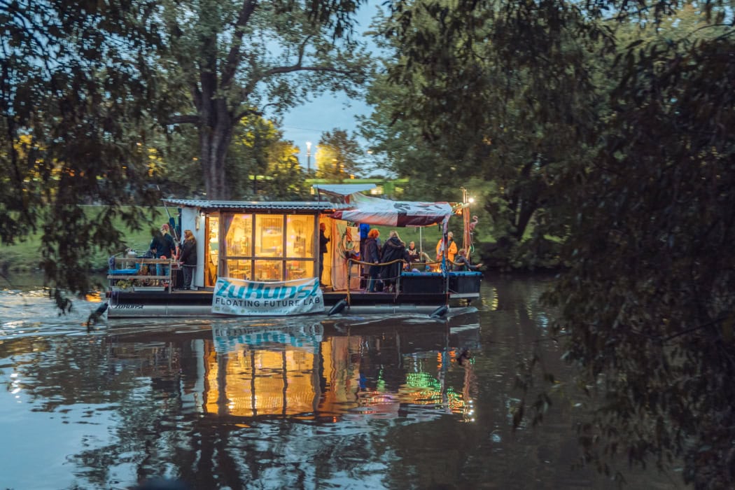 Im abendlichen Halbdunkel sieht man ein von innen in gelbem Licht erleuchtetes Hausboot einen Fluss entlang fahren.