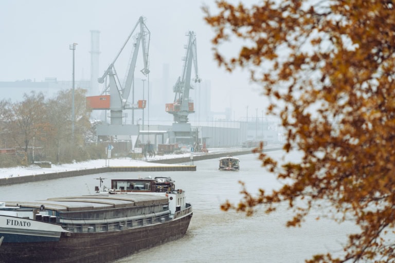 In einer schneebedeckten Hafenlandschaft sieht man einen Fluss, auf dem aus dem linken unteren Bildrand ein Frachtschiff kommt. Dem Frachtschiff gegenüber schwimmt ein fast schon winzig wirkendes Hausboot. Rechts im Bild sind unscharfe, braune Blätter im Vordergrund. Im Hintergrund erkännt man zwei große Hafenkräne und ein Industriegebiet.