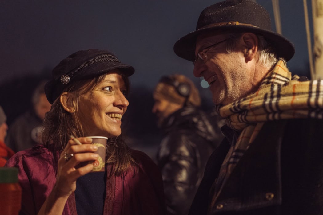 Eine Frau (links) und ein Mann (Rechts), in der Portraitperspektive fotografiert, unterhalten sich. Die Frau, deren Gesicht erleuchtet ist, trägt eine rote Bluse und eine schwarze Mütze, sie hält einen Pappbecher in der Hand. Sie lacht und blickt nach rechts zu dem Mann, dessen Gesicht im Schatten ist, auch er lacht. Er trägt einen Hut und einen karierten Schal. Im Hintergrund sind unscharf weitere Personen zu sehen.