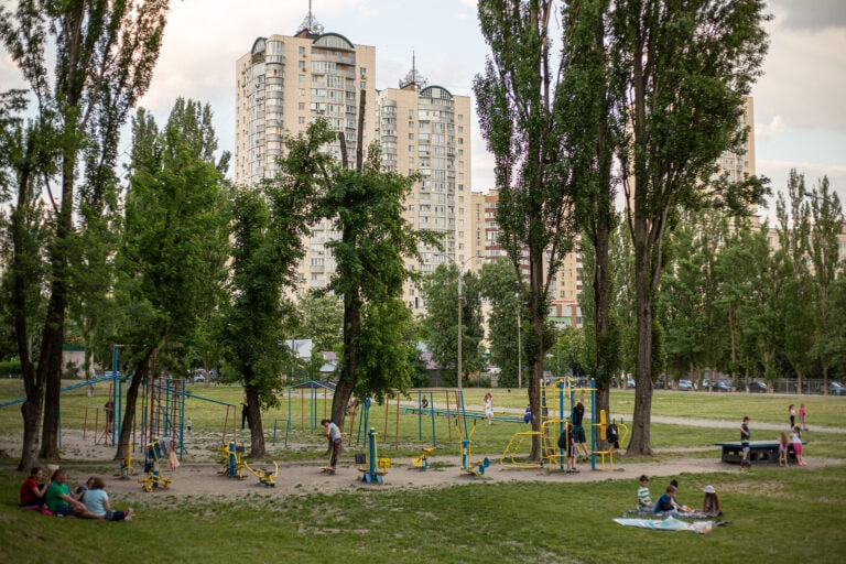 Park in Kiev, im Hintergrund Hochhäuser, im Vordergrund ein Spielplatz mit Familien und Kindern. Fotografiert von Tetiana Cherniavska im Mai 2023.