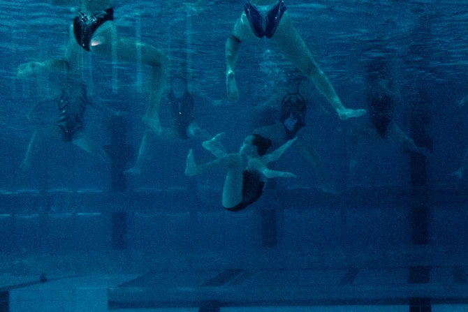 Mehrere Synchronschwimmerinnen befinden sich während der Aufführung einer Choreografie in einem Schwimmbecken. Die Synchronschwimmerinnen sind teilweise oder komplett Unterwasser zu sehen.