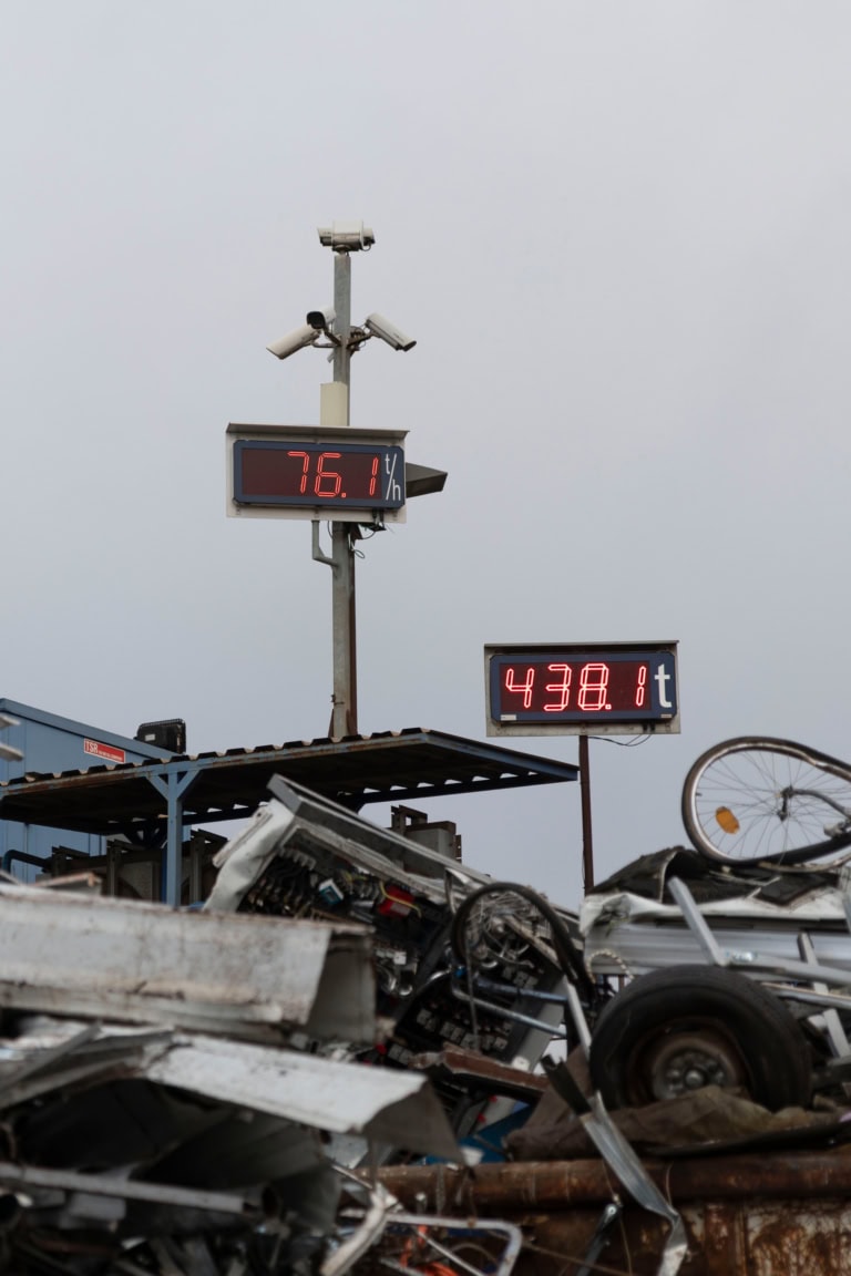 Digitale Anzeigetafeln auf einem Schrottplatz zeigen 76,1 t/h und 438,8 t über einem Haufen aus Metall- und Fahrzeugteilen