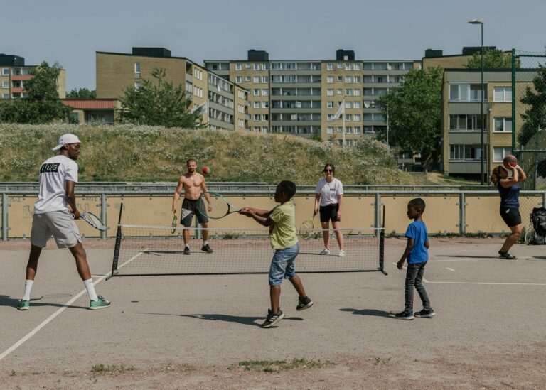 Eine Gruppe von Menschen, darunter Kinder und Erwachsene, spielt Tennis auf einem städtischen Platz in Malmö-Rosengard, umgeben von Wohngebäuden. Die Szene zeigt eine lebendige Gemeinschaftsaktivität, ideal für diejenigen, die Fotografie Studieren und das urbane Leben dokumentieren möchten. Dieses Bild wurde von einer Fotografie-Studentin aufgenommen.