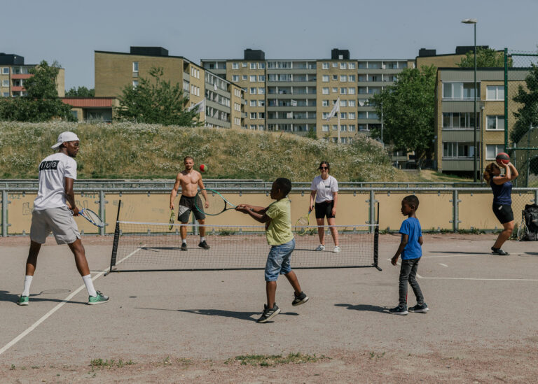 Jugendliche spielen im Viertel Rosengard in Malmö Tennis auf einem Sportplatz, im Hintergrund Wohnblocks aus den 1960er Jahren.