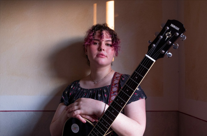 Musician Nina Freckles with pink curls holds a black guitar against a pale wall – self-empowerment through music