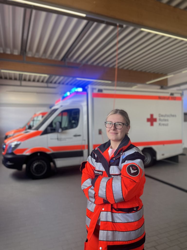 Paula S. gehört zur Gen-Z. Sie steht lächelnd vor mehreren Rettungswägen in einer Halle und hat die Arme verschränkt. Sie trägt eine Uniform des Deutschen Roten Kreuz.