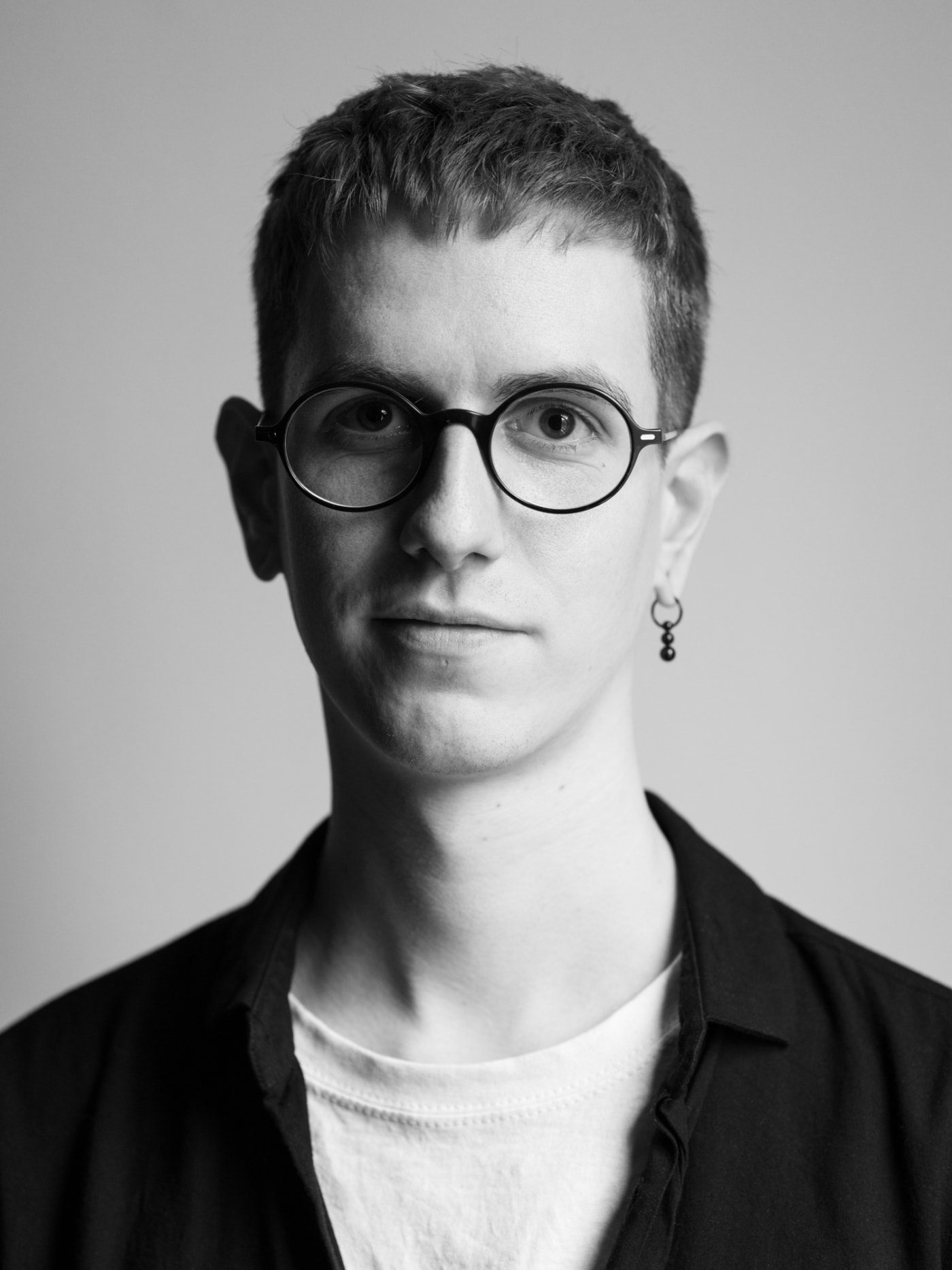 Markus Heft posiert für ein Portrait in Schwarz Weiß. Er schaut direkt in die Kamera. Markus Heft trägt eine runde Brille und einen hängenden Ohrring.