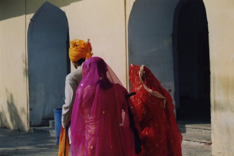 Rückansicht von zwei Frauen in farbenfrohen Saris (Pink und Rot) und einem Mann mit gelbem Turban, vor einem historischen Gebäude mit Bögen.