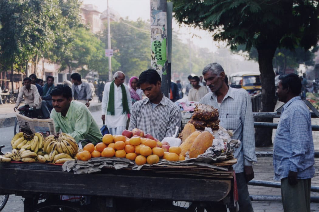 Straßenszene in Indien mit Obst- und Gemüsestand, auf dem Bananen, Orangen und Papayas angeboten werden, während Männer in traditioneller Kleidung dahinter stehen und Zeitung lesen und die Umgebung lebhaft ist.