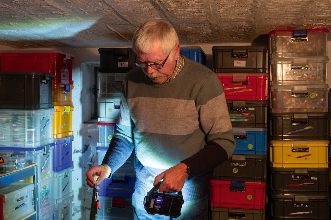 Kugelschreiber-Sammler Heinz-Hermann Bendig steht in einem dunklem Raum, in welchem sich bunte Plastikkisten bis zur Decke stapeln. Die Kisten sind gefüllt mit Kugelschreibern. In der Hand hält er eine Taschenlampe und beleuchtet einen Kuli.