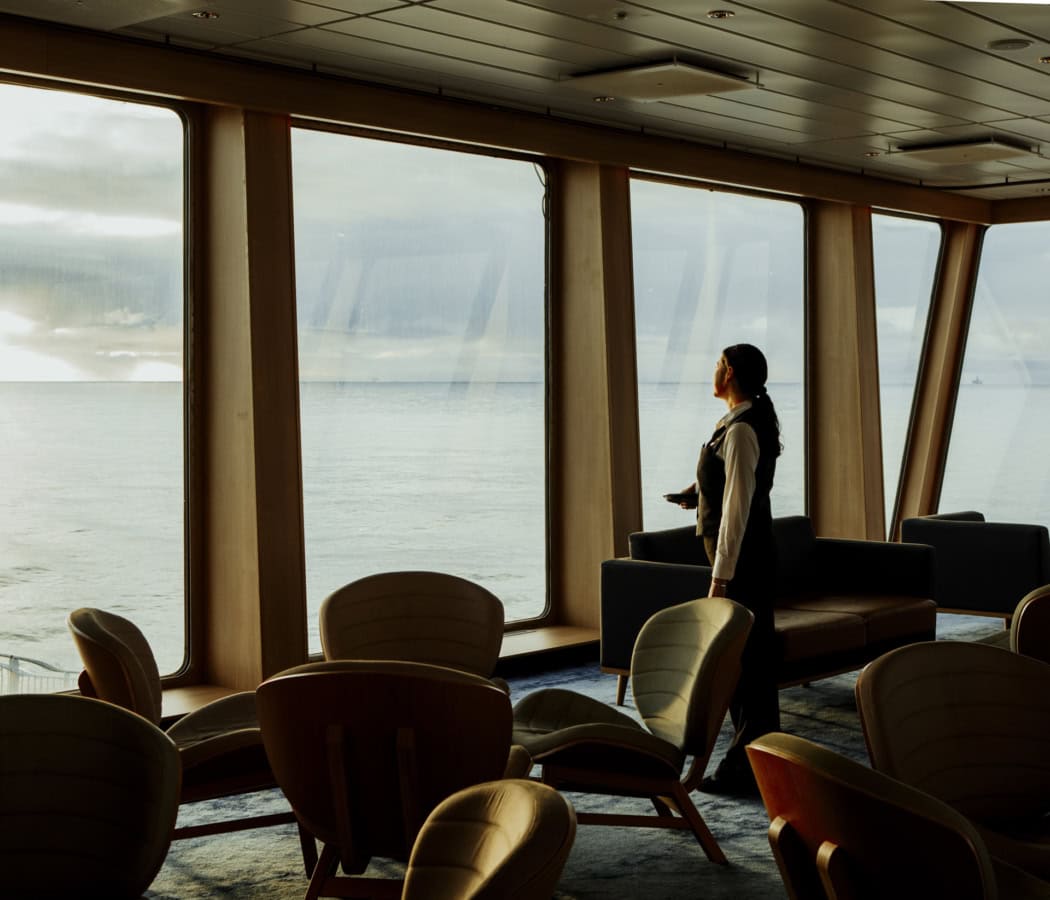 Ein Besatzungsmitglied steht im Innenraum der MS Norröna, wo mehrere leere Tische und Stühle vor großen Fenstern stehen. Das sanfte Licht des frühen Abends schafft eine träumerische Atmosphäre.