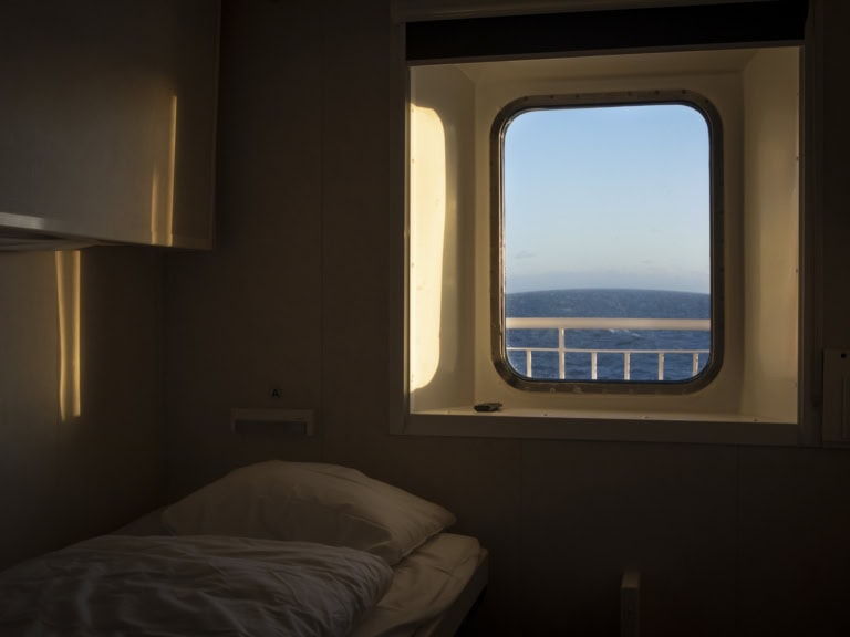 Eine Kabine der MS Norröna ist mit einem Bett ausgestattet. Man hat durch ein kleines Fenster den Blick aufs Meer. Die Abendsonne scheint in den Raum und sorgt für eine gemütliche Atmosphäre.