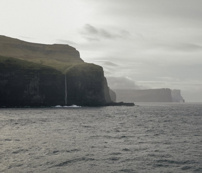 Die MS Norröna passiert die raue Landschaft mit satten grünen Wiesen und Wasserfällen der Färöer Inseln. In der Ferne regnet es.