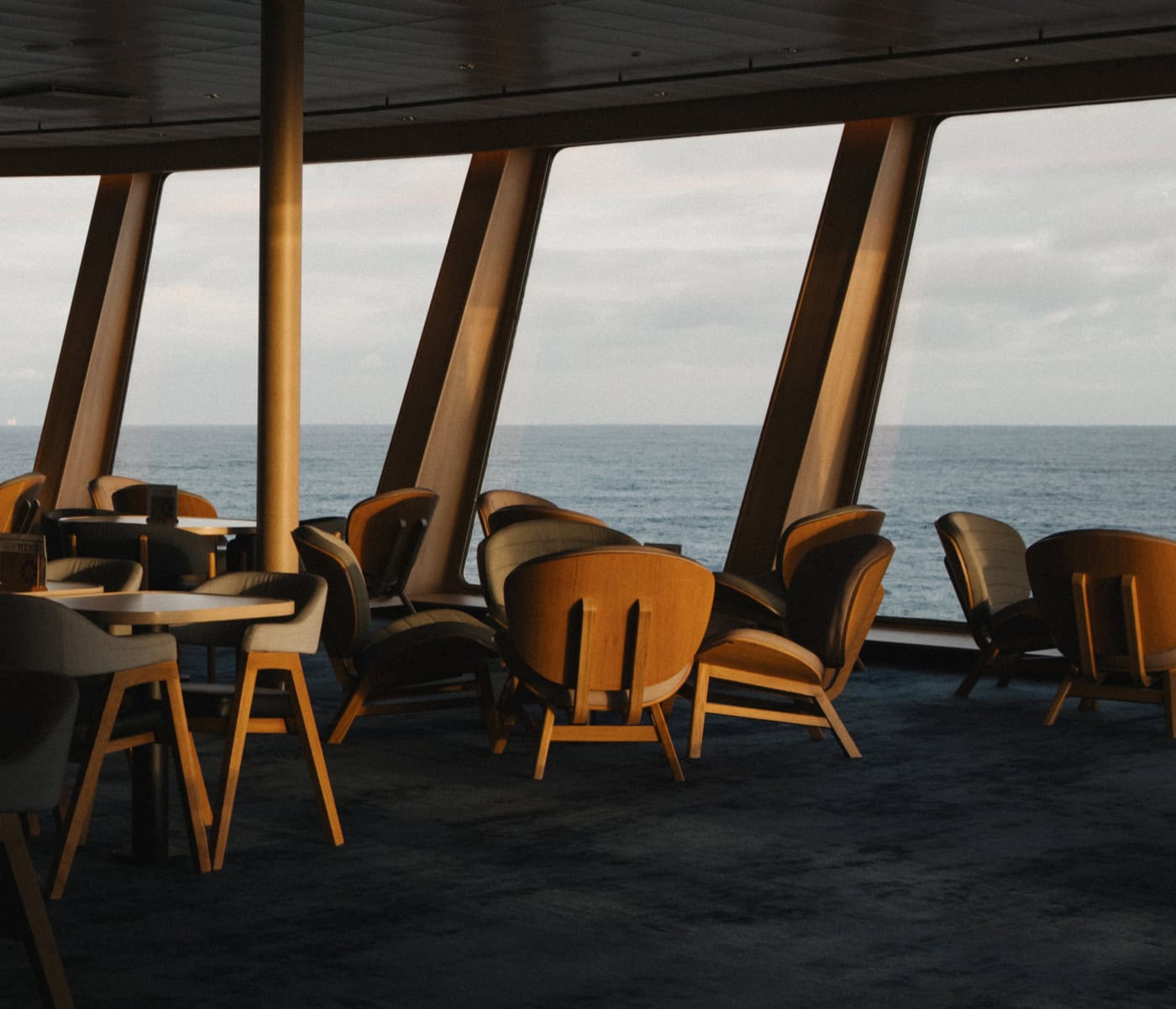 Das Bild zeigt den Innenbereich der MS Norröna mit mehreren leeren Tischen und Stühlen vor großen Fenstern, die einen weiten Blick auf das Meer bieten. Das sanfte Licht des frühen Abends erzeugt eine gemütliche Atmosphäre.