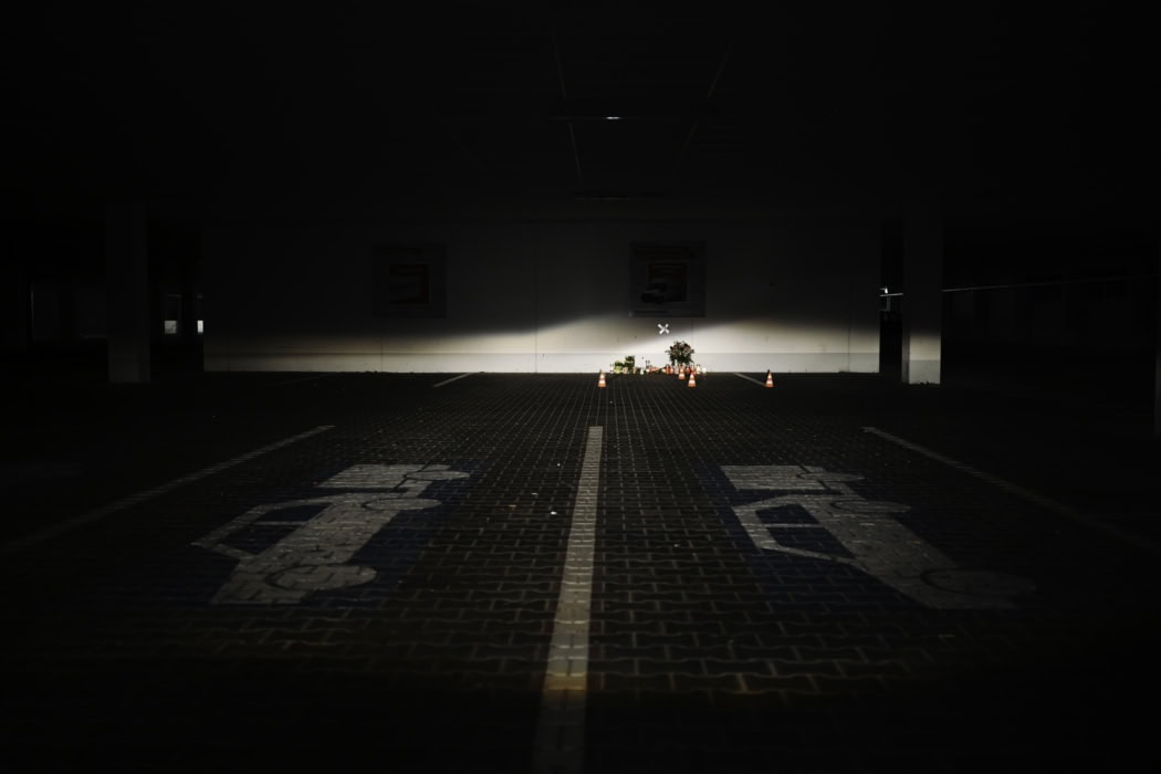 Das Bild zeigt eine Tiefgarage bei Nacht. Im Zentrum des Bildes, beleuchtet von einer Taschenlampe, befindet sich ein kleiner Gedenkort mit Blumen, Kerzen und Absperrkegeln. Auf dem Boden sind Parkflächenmarkierungen mit der Umrissdarstellung von Autos zu sehen. Das Bild zeigt einen Tatort eines Femizids in Regensburg. Parkplatz Baummarkt Toom