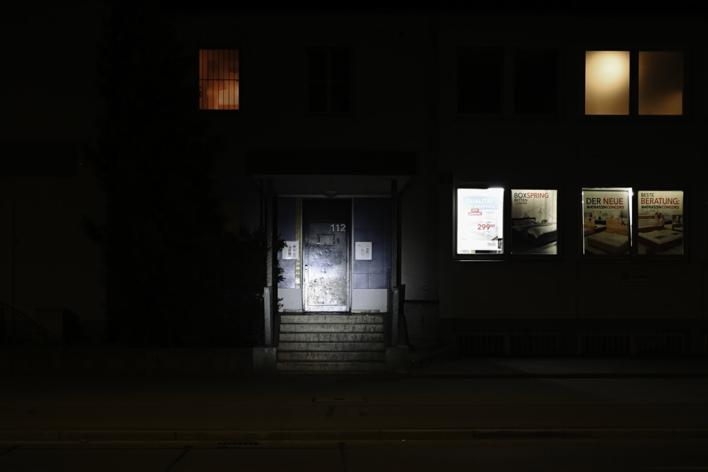 Das Bild zeigt nachts ein Gebäude mit einer beleuchteten Eingangstür mit der Nummer 112. Im Erdgeschoss befinden sich Schaufenster mit Werbung für Betten und Matratzen. Ein Fenster im oberen Stockwerk leuchtet rot, zwei andere orange. Das Bild zeigt einen Tatort eines Femizids in Regensburg. Landshuter Straße 112.