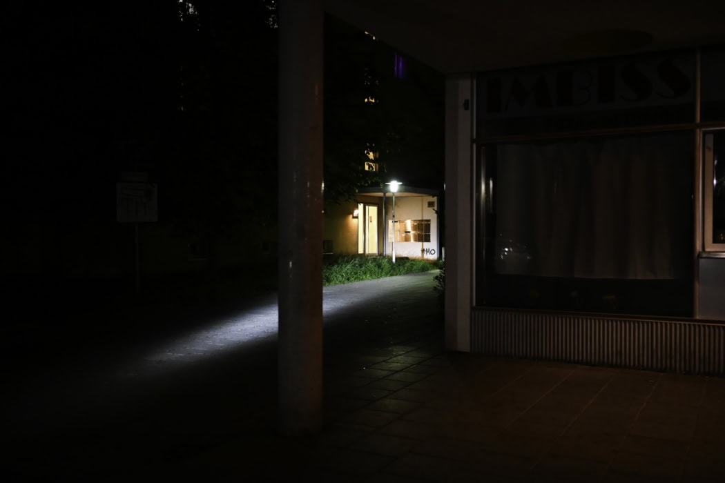 Das Bild zeigt eine dunkle, nächtliche Szene. Im Vordergrund steht eine dicke Säule, die den Blick auf einen schwach beleuchteten Gehweg teilt, der zu einem beleuchteten Gebäude führt. Rechts im Bild ist ein Gebäude mit eeinem Schaufenster, das mit einem Vorhang verdeckt. Das Bild zeigt einen Tatort eines Femizids in Regensburg. Humboldtstraße 40