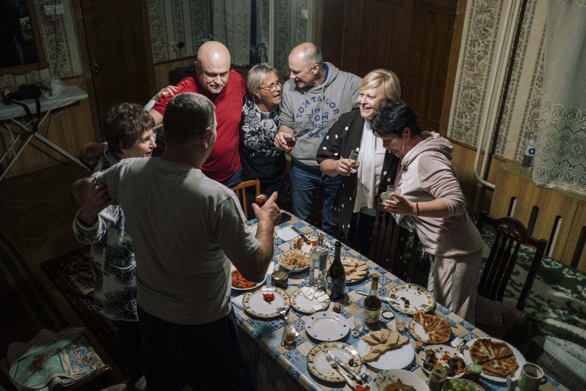 Eine Gruppe von Menschen steht lachend und fröhlich um einen Esstisch herum, der mit verschiedenen Speisen und Getränken gedeckt ist. Sie wirken vertraut und glücklich, als ob sie ein Familientreffen oder eine Feier genießen. Die Szene strahlt Wärme und Gemeinschaft aus und vermittelt ein starkes Heimatgefühl.