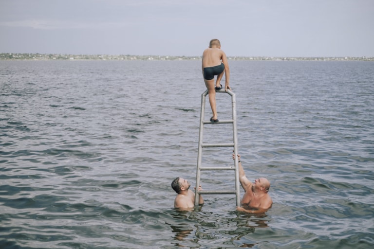 Zwei Männer und ein Kind genießen gemeinsam einen Tag im Wasser. Das Kind klettert eine Metallleiter hinauf, während die Männer es von unten unterstützen und beobachten. Im Hintergrund ist eine weite Wasserfläche zu sehen, die bis zum Horizont reicht.