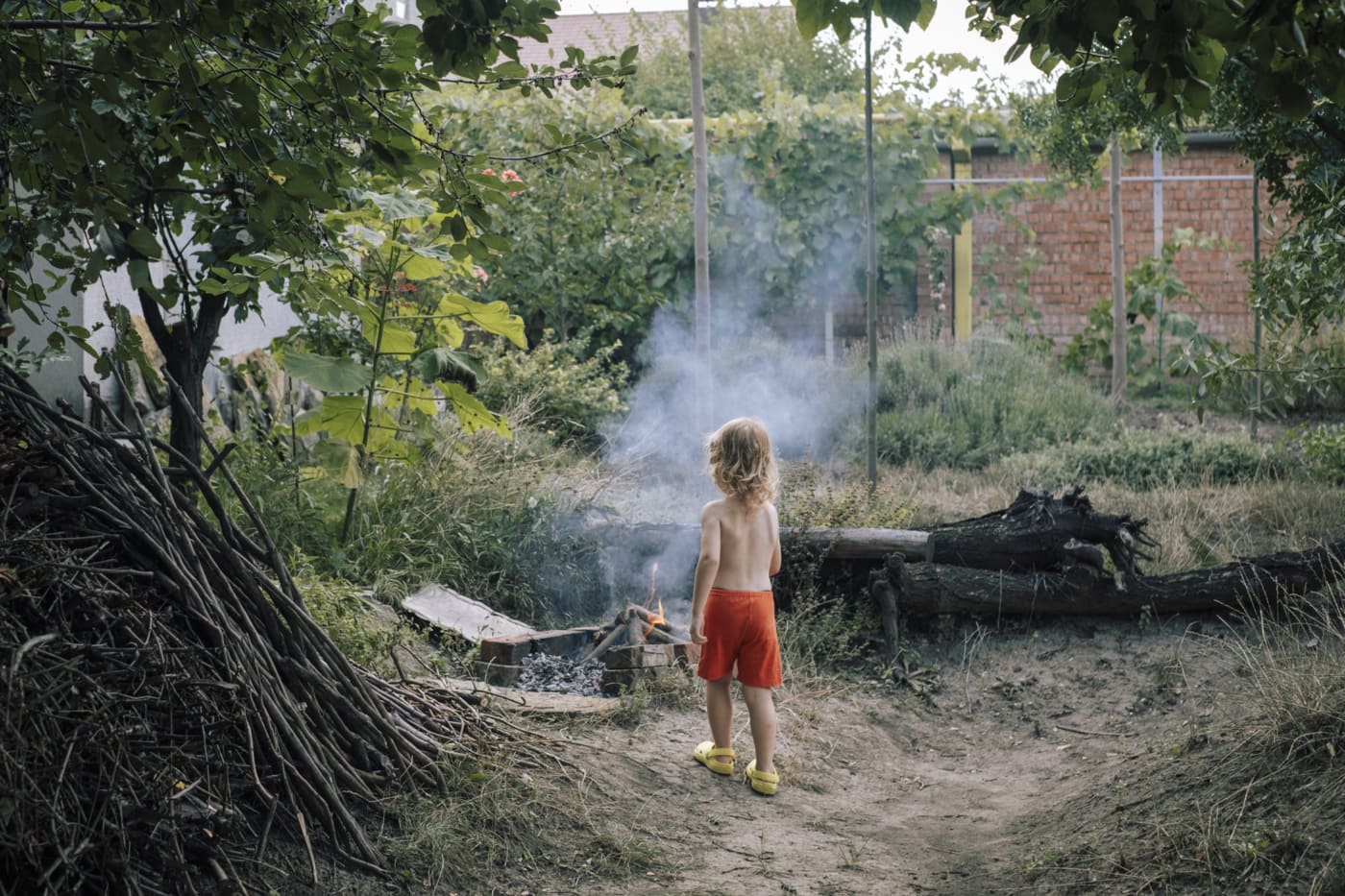 Ein kleines Kind mit langen, blonden Haaren und roten Shorts steht mit dem Rücken zur Kamera und schaut auf ein kleines Feuer in einem Garten. Der Garten ist dicht bewachsen mit grünen Pflanzen und Bäumen. Im Hintergrund ist ein Zaun und ein Haus zu sehen.