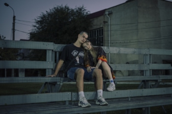 Ein junges Paar sitzt auf einer leeren Tribüne bei Abenddämmerung, das Mädchen lehnt sich an die Schulter des Jungen und beide tragen lässige Kleidung. Der Hintergrund zeigt eine urbane Umgebung mit Bäumen und einem Gebäude.