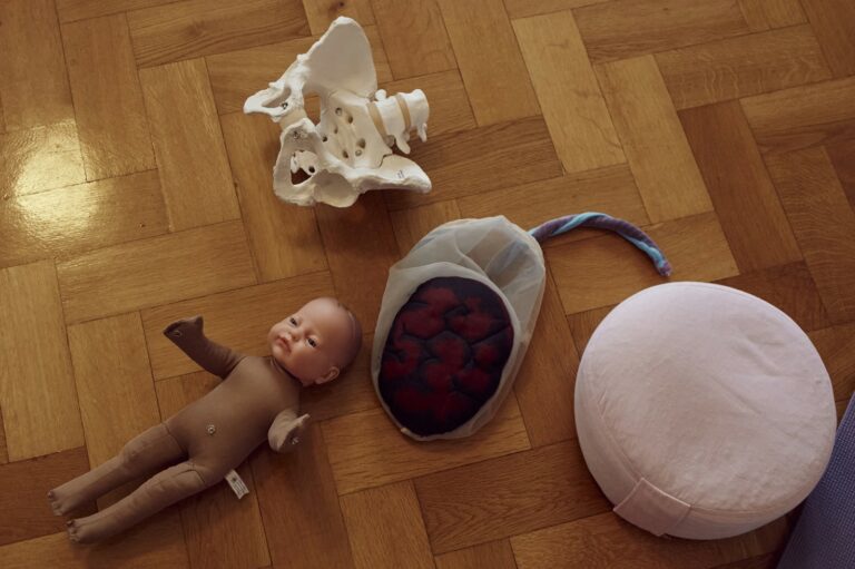 Auf dem Boden liegen eine Puppe, die Nachbildung eines Beckens und eine Plazenta mit Fruchtblase und Nabelschnur aus Stoff. Die Gegenstände veranschaulichen im Geburtsvorbereitungskurs den Geburtsverlauf.