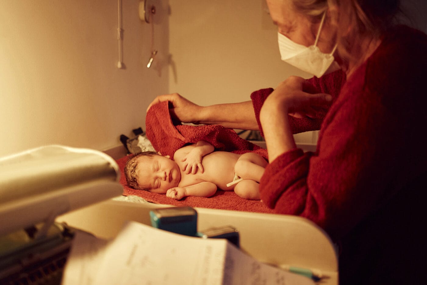 A newborn baby lies on a changing table. The midwife stands in front of the baby, drying it with a red towel.