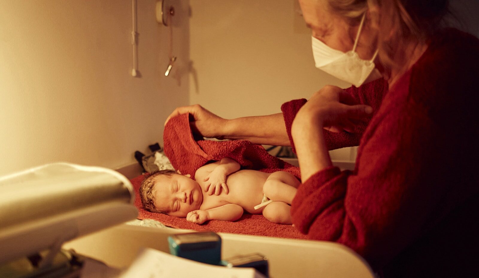 Ein Neugeborenes liegt auf einem Wickeltisch. Die Hebamme steht vor dem Kind und trocknet es mit einem roten Handtuch ab.