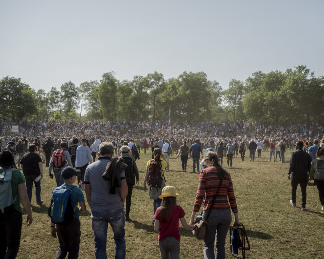 Eine große Menschenmenge versammelt sich auf einem Feld während des Dülmener Wildpferdefangs. Im Hintergrund sitzen Zuschauer auf Tribünen, umgeben von grünen Bäumen und unter einem klaren, blauen Himmel. Die Veranstaltung zieht zahlreiche Besucher an, die gespannt das Geschehen verfolgen.