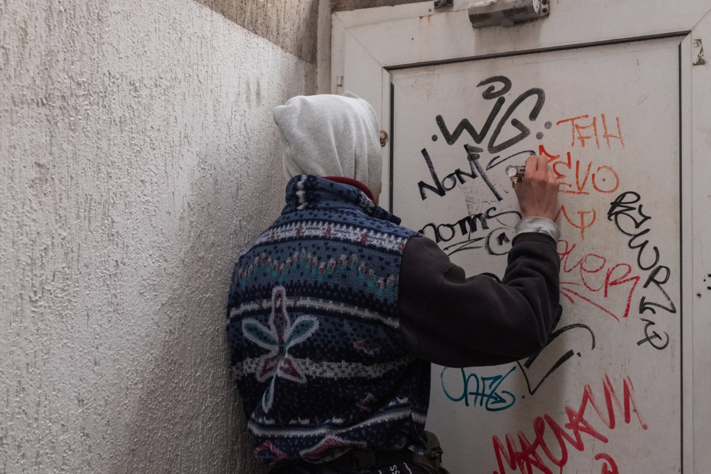 Person mit Kapuzenpullover und gemusterter Weste schreibt Graffiti auf eine weiße Tür in einem engen Durchgang