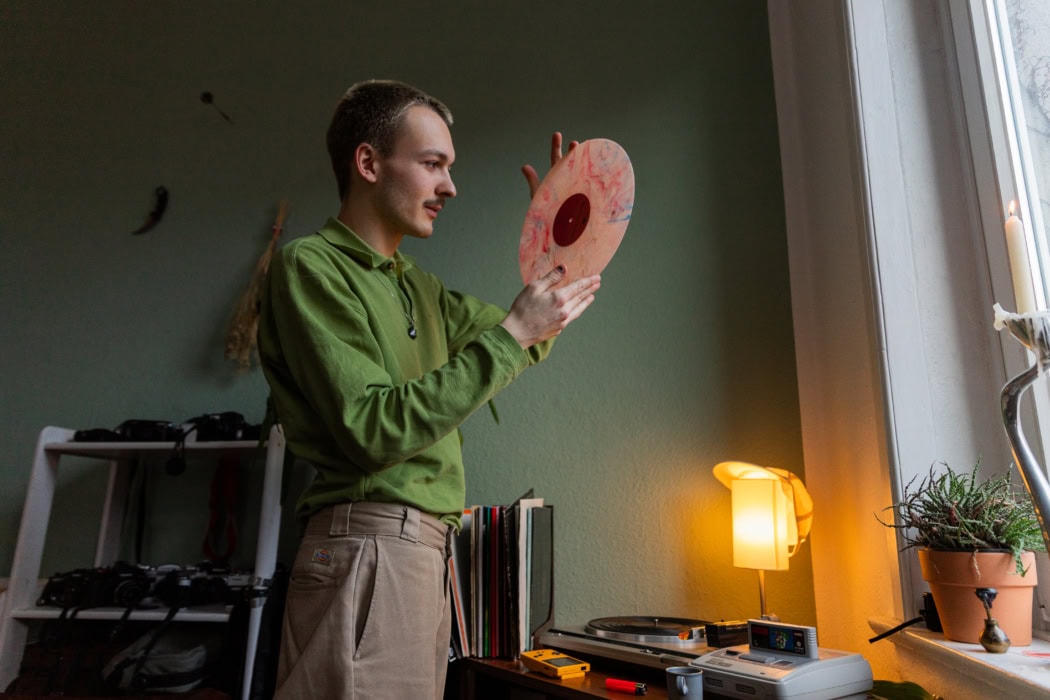 Ein junger Mann hält eine Schallplatte in der Hand. Er steht in einem Raum vor einem Fenster neben einem kleinen Tisch, auf dem ein Plattenspieler steht. Hinter dem Tisch steht eine Lampe. Am Fensterrand steht eine grüne Pflanze. Der junge Mann trägt einen grünen Wollpullover und eine beige Hose.