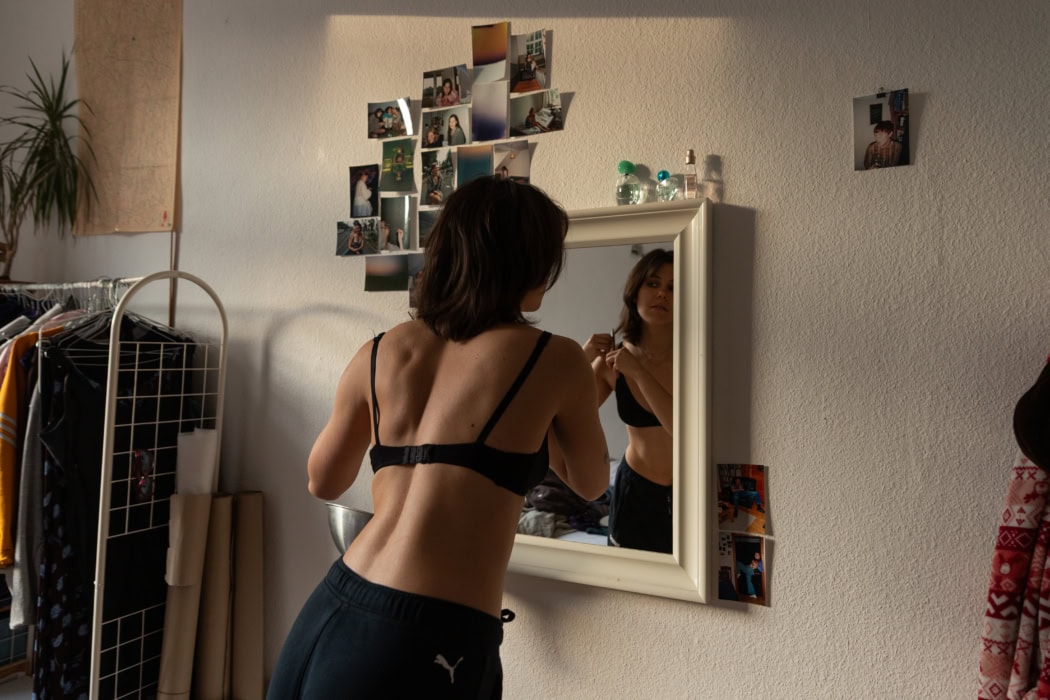 Eine junge Frau steht vor einem Spiegel und schneidet sich die Haare. Sie befindet sich in einem Zimmer. An der Wand hängen eine Reihe von Fotos, neben dem Mädchen Kleider, eine Pflanze und eine Landkarte.