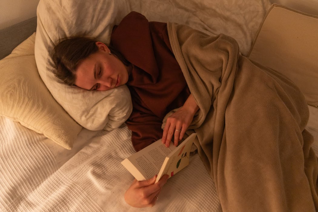 Eine junge Frau liegt auf dem Bett und liest in einem Buch. Sie ist in eine Decke gehüllt. Sie trägt einen weinroten Pullover. Auf dem Bett liegen Kissen.