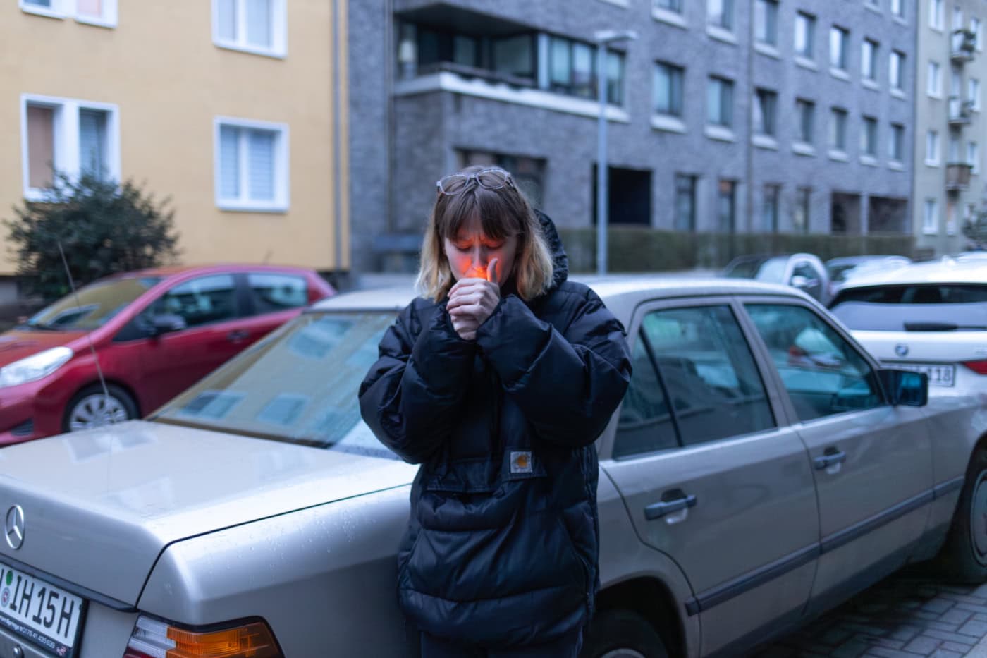 Junge Frau in dunklem Wintermantel zündet eine Zigarette an, steht vor einem geparkten silbernen Mercedes in einer Wohnstraße